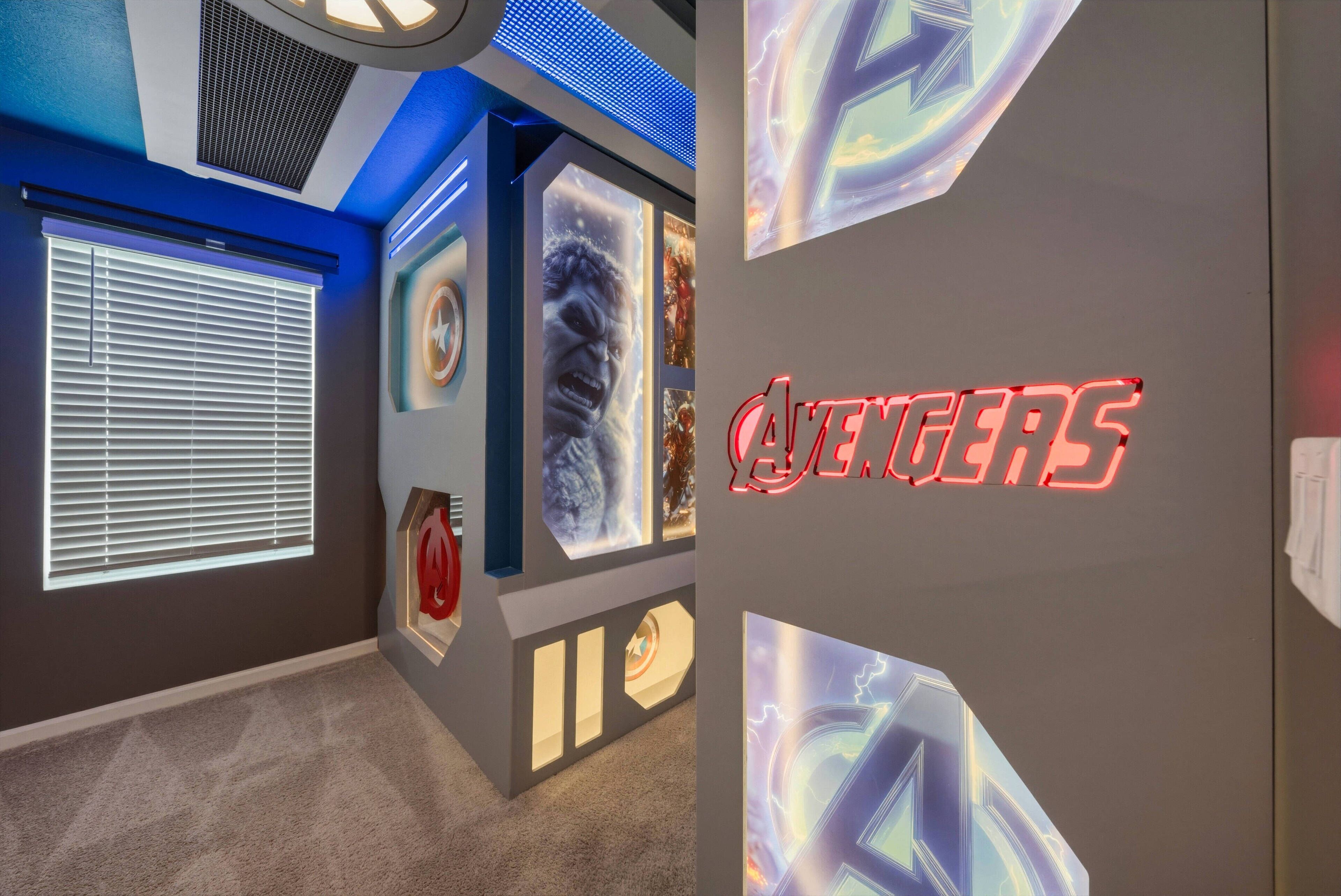 Een kamer met een grote muurschildering met een thema van de Avengers, een raam met zonwering en een ventilatie rooster in het plafond.