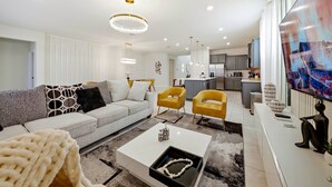 Een moderne woonkamer met een bank, salontafel en twee gele stoelen. Achtergrond is een keuken met een koelkast en kasten.