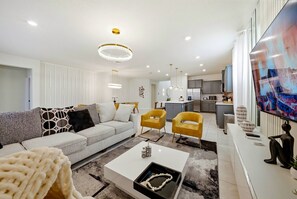 A modern living room with a sofa, coffee table, and two yellow chairs. There is a kitchen in the background with a refrigerator and cabinets. - 8BR house at Windsor Cay Resort (Clermont)