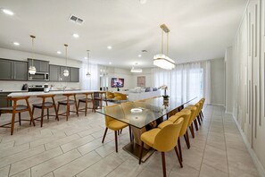 A modern kitchen with grey cabinets, a central island, and a dining area with yellow chairs and a glass table. - 8BR house at Windsor Cay Resort (Clermont)