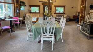 Un espace repas avec une longue table, des chaises blanches et une nappe verte. On y trouve des fenêtres avec des rideaux, un ventilateur de plafond et des guirlandes lumineuses.