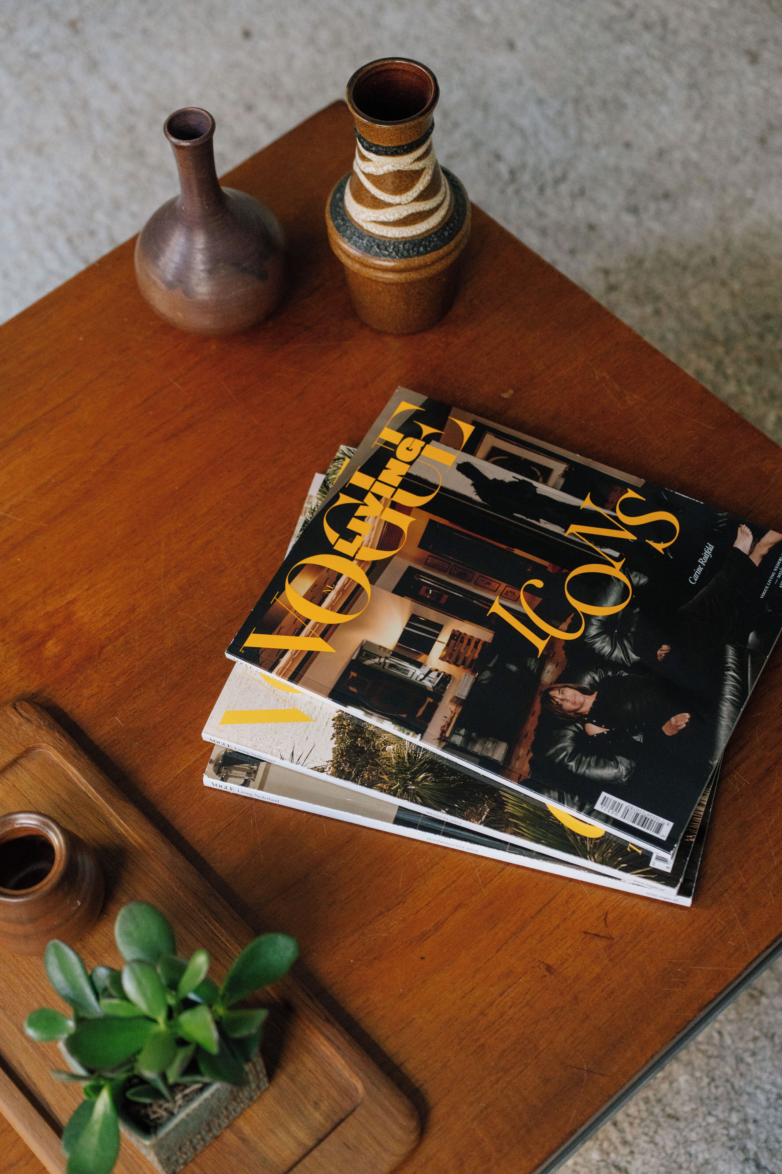 A stack of magazines on a wooden table, accompanied by a small potted plant and two ceramic vases.