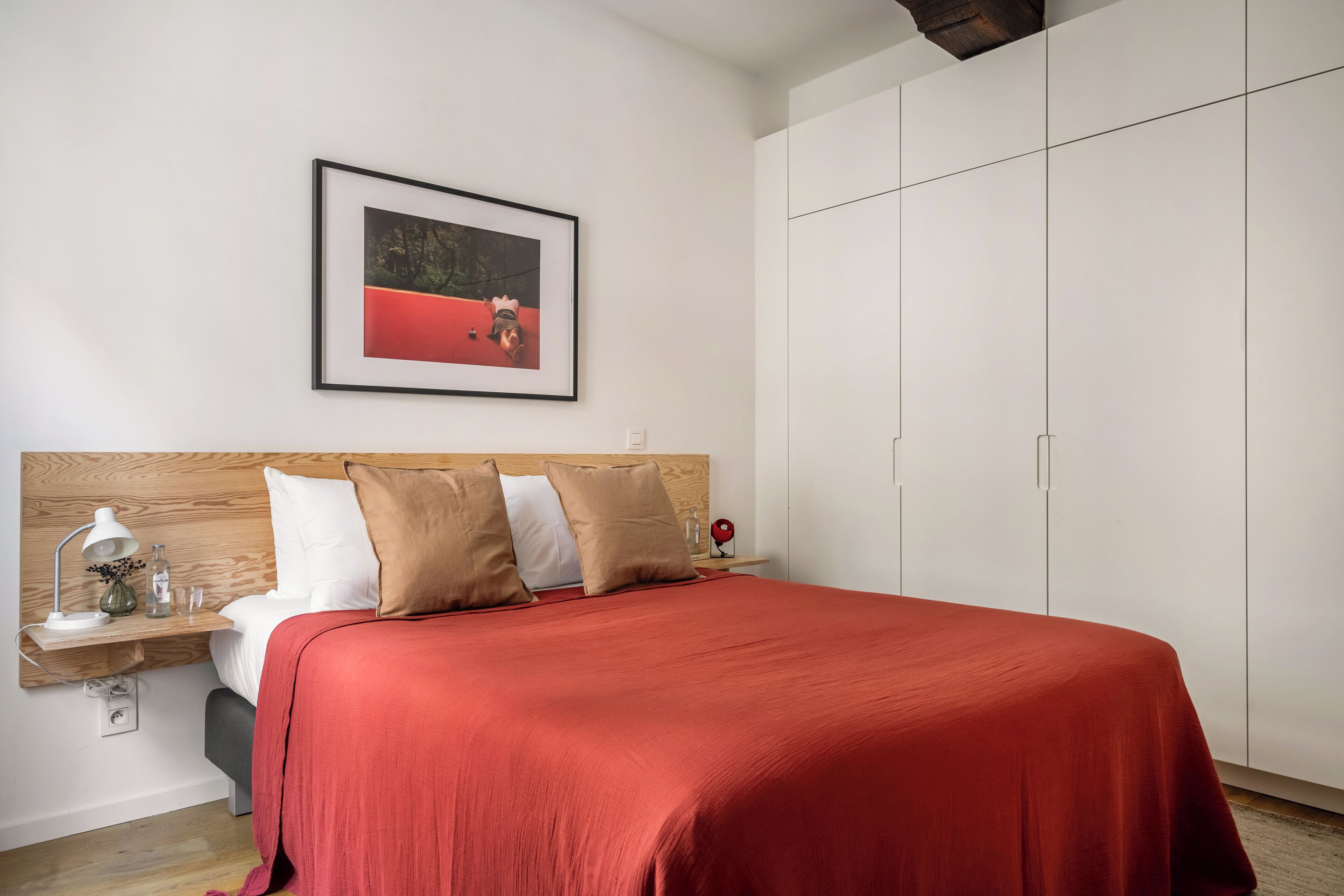 Een slaapkamer met een houten hoofdbord, een rode spreideken, een nachtkastje met een lamp, en een ingelijst beeld aan de muur.