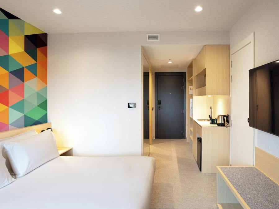 A hotel room with a bed, a TV, a kitchenette, and a colorful geometric wall design.