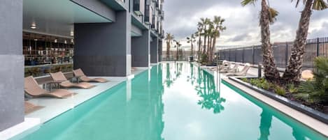 A modern hotel with a long swimming pool, lounge chairs, and palm trees.