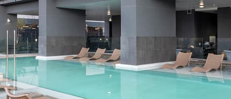 A modern indoor pool with lounge chairs, a tiled floor, and a building with balconies.