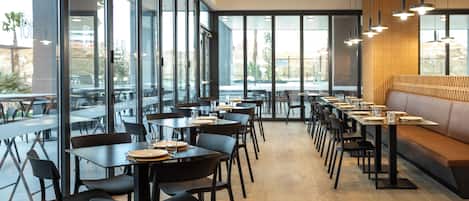 Un intérieur de restaurant moderne avec de longues tables, des chaises noires et un espace détente avec des bancs en bois.