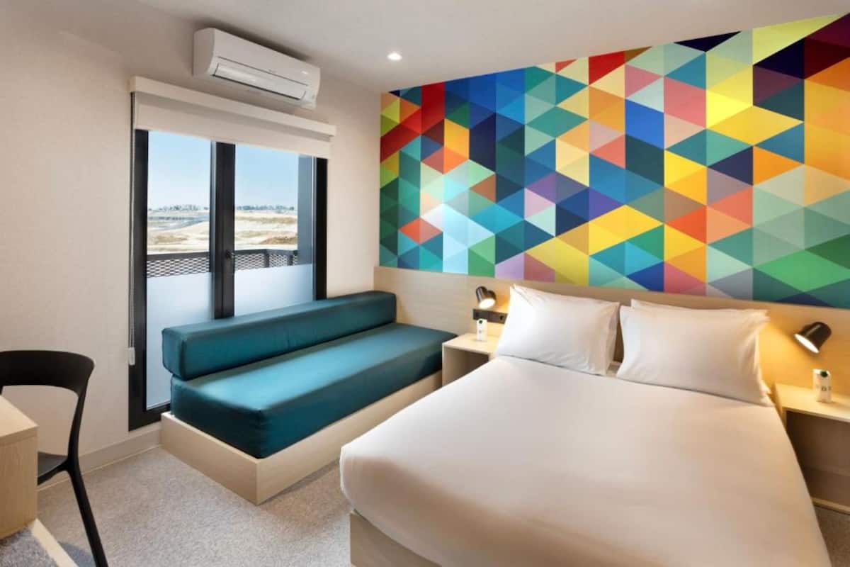 A hotel room with a bed, a bench, a desk, and a colorful geometric wall art.