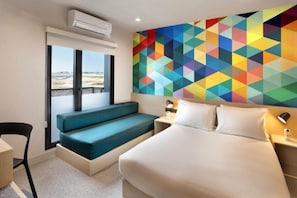Quarto de hotel com uma cama, um banco, uma escrivaninha e uma obra de arte geométrica colorida na parede.