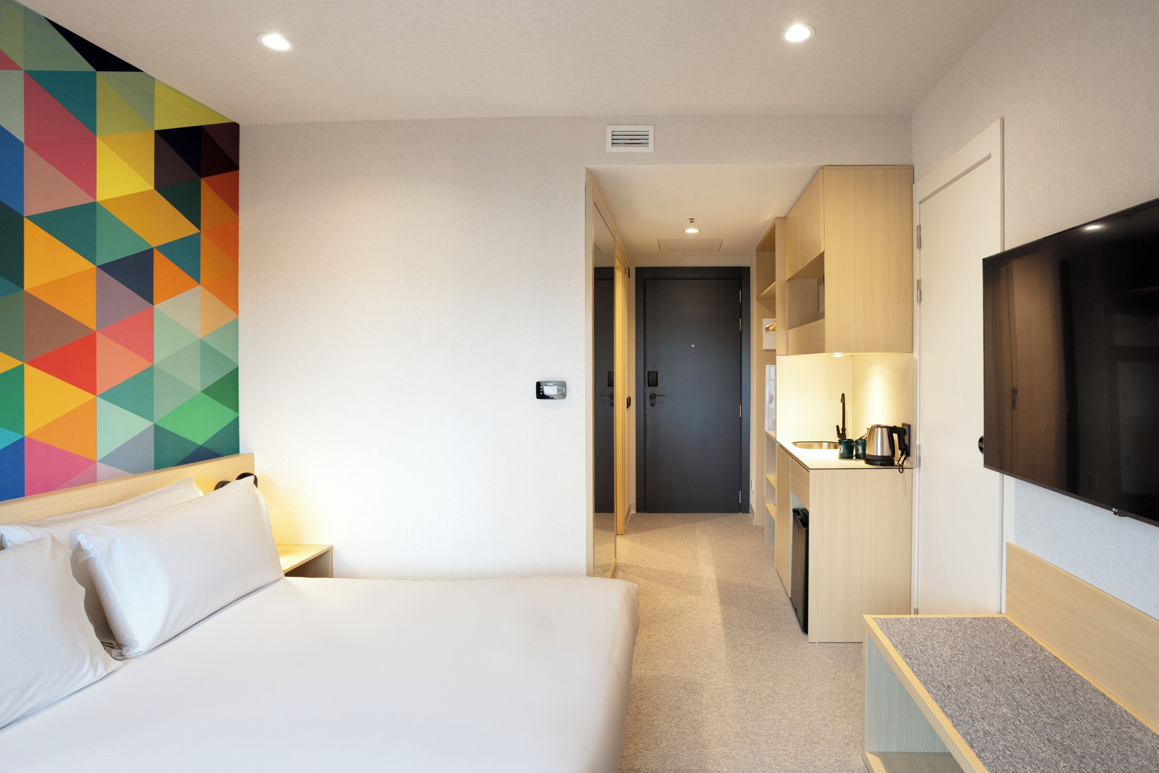 A hotel room with a bed, a TV, a kitchenette, and a colorful geometric wall design.