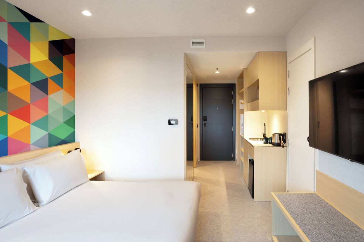 A hotel room with a bed, a TV, a kitchenette, and a colorful geometric wall design.