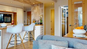 A modern kitchen with white chairs, a grey sofa, and a wooden dining table with a bowl of fruit.