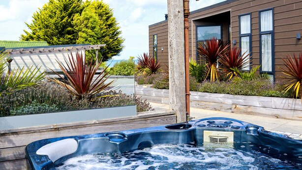 A hot tub in a landscaped area with a wooden deck, surrounded by plants and a modern building with large windows.