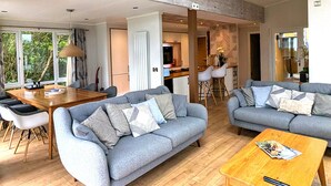 A spacious living area with grey sofas, a wooden coffee table, and a dining table with chairs. There is a kitchen area in the background with a bar and stools.