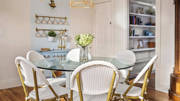 Un comedor con una mesa de vidrio rodeada de sillas de mimbre blanco con detalles dorados, un jarrón con flores y un estante de pared con objetos decorativos.