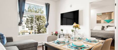 A modern living room with a dining area, a flat-screen TV, and a bed with a geometric-patterned headboard.