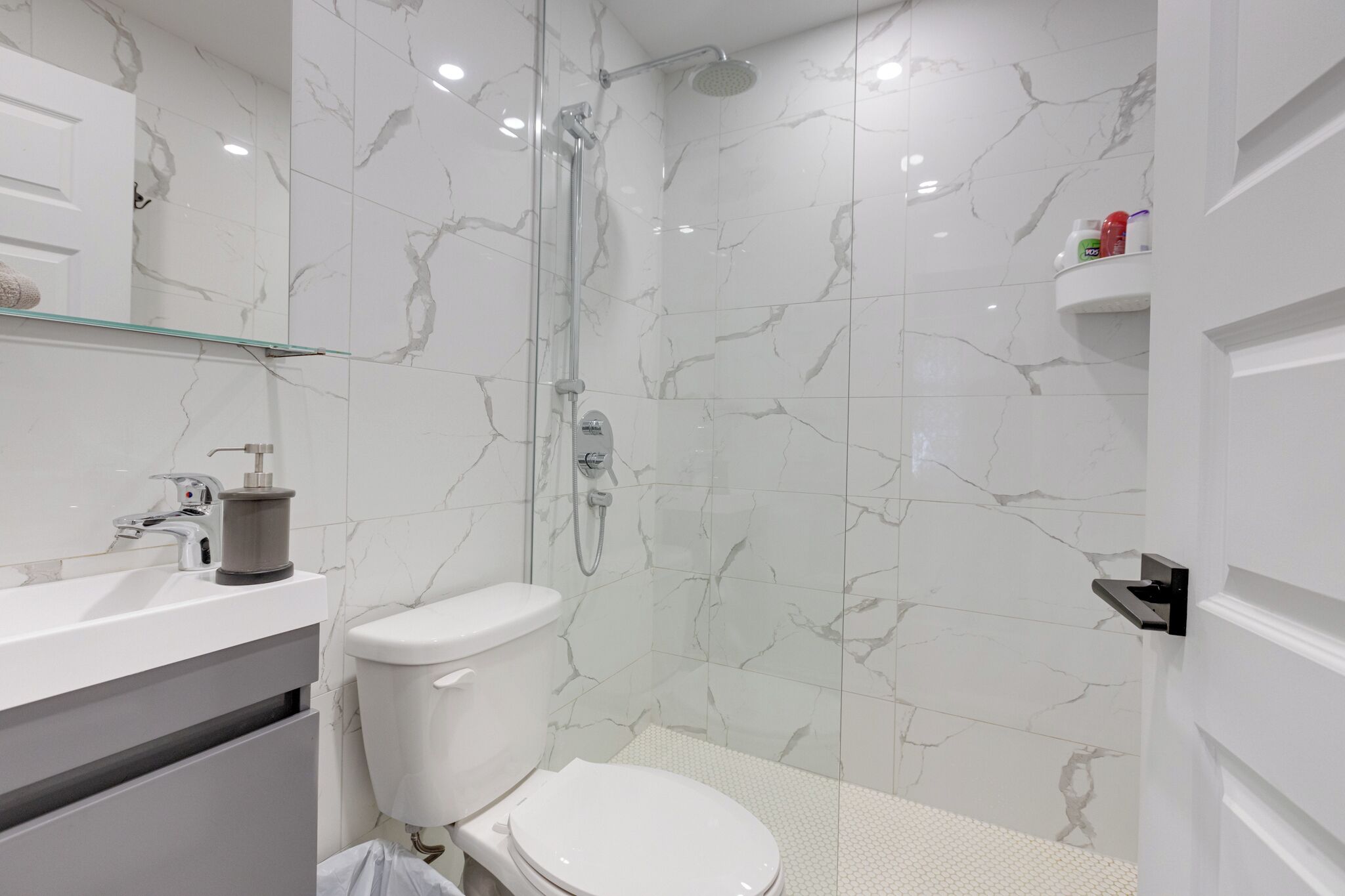 A modern bathroom with a glass shower enclosure, white marble tiles, a white toilet, and a white sink with a soap dispenser.