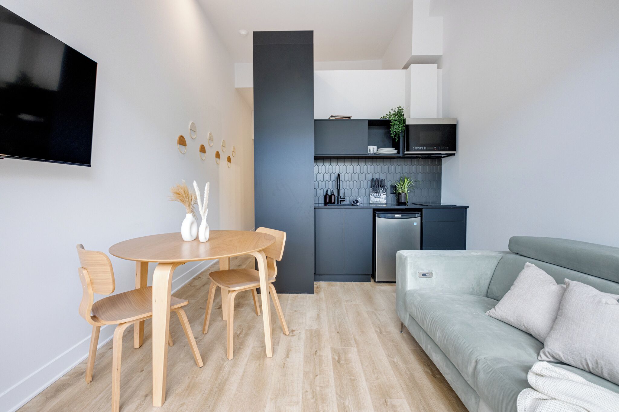 A modern living room with a grey sofa, wooden dining table, and chairs. A flat-screen TV is mounted on the wall.
