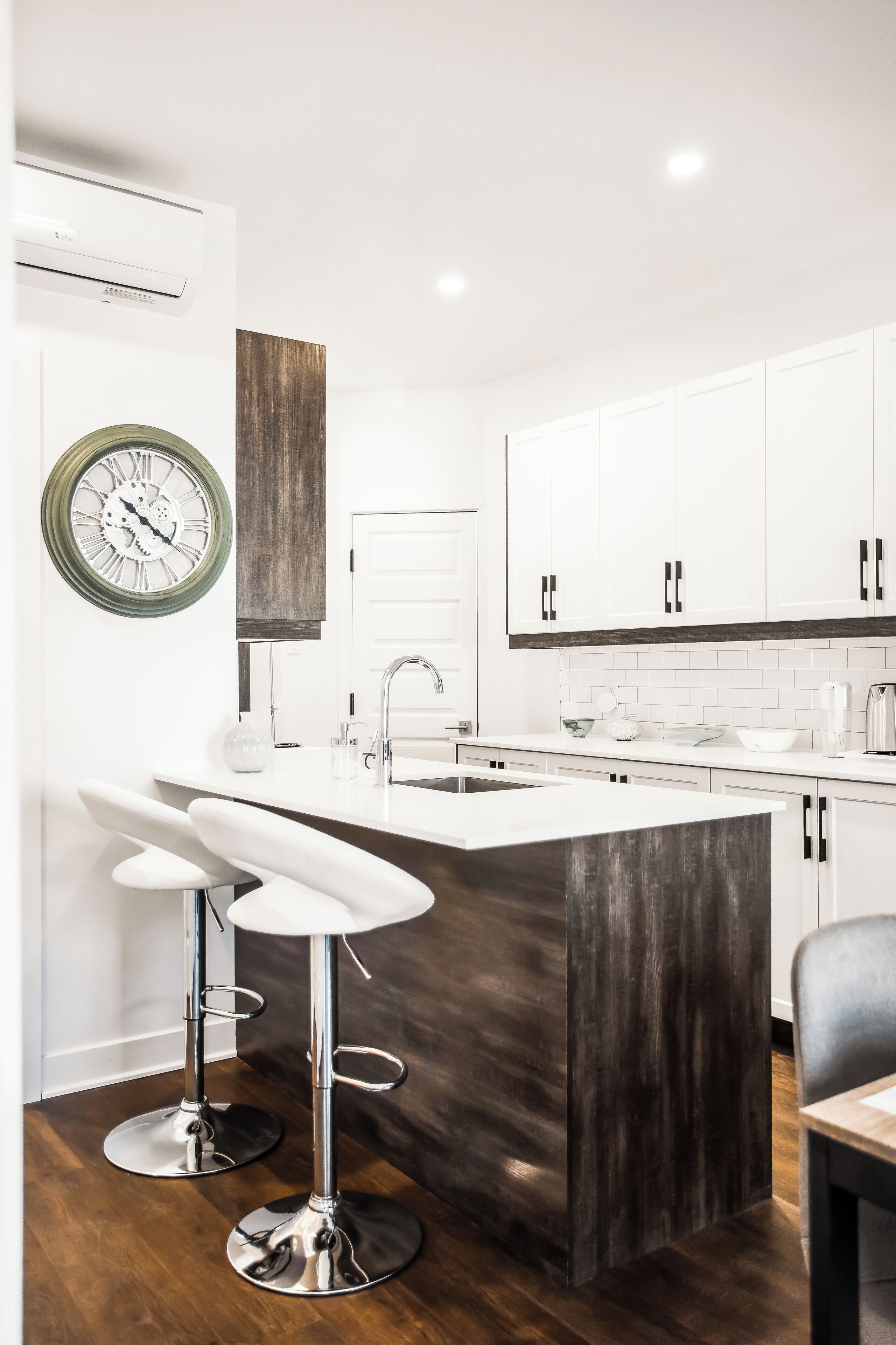 Una cocina moderna con armarios blancos, una isla de madera, taburetes de barra de acero inoxidable y un reloj fijado a la pared.