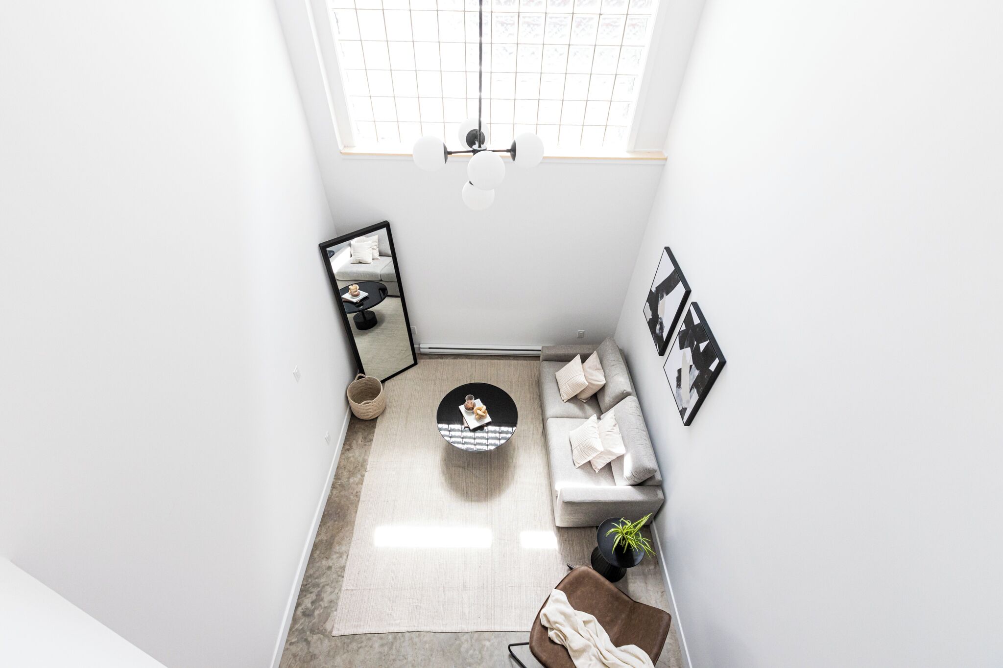 Une pièce moderne et minimaliste, dotée d’une grande fenêtre, d’une table basse ronde, d’un canapé, d’un miroir et d’une petite plante.