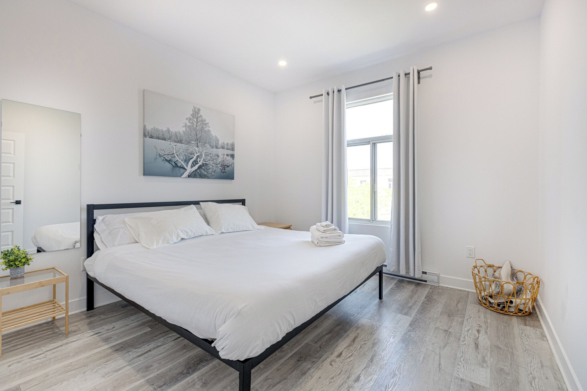 A modern bedroom with a large bed, a bedside table with a plant, a wall-mounted painting, and a window with curtains.