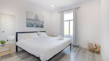 Een moderne slaapkamer met een groot bed, een nachtkastje met een plant, een aan de muur hangend schilderij en een raam met gordijnen.