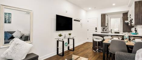 A modern living room with a flat-screen TV, a glass coffee table, and a dining area with a wooden table and chairs.