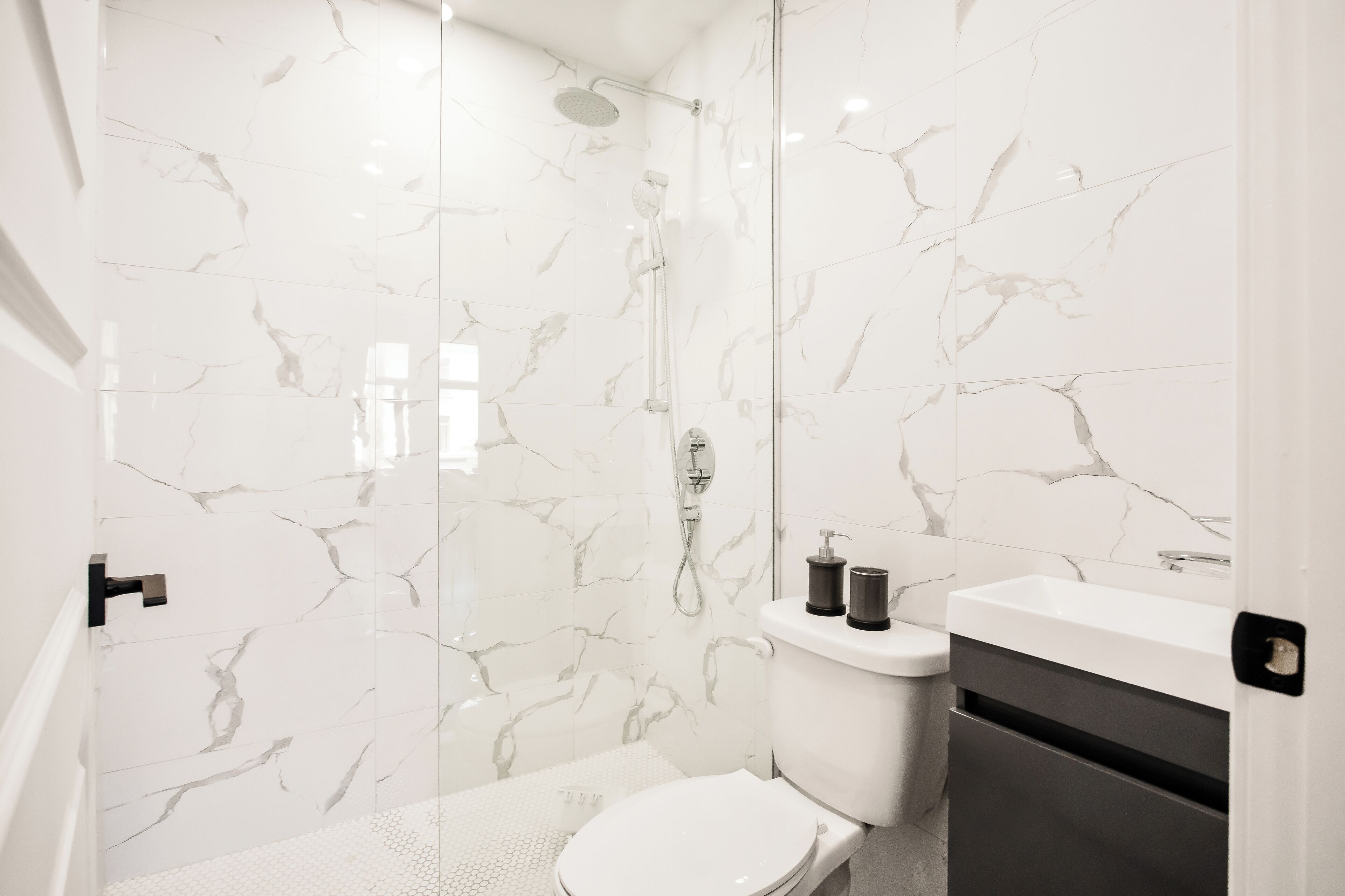 Een moderne badkamer met een douchecabine van glas, witte marmeren wanden, een wit toilet en een donkere wastafel.