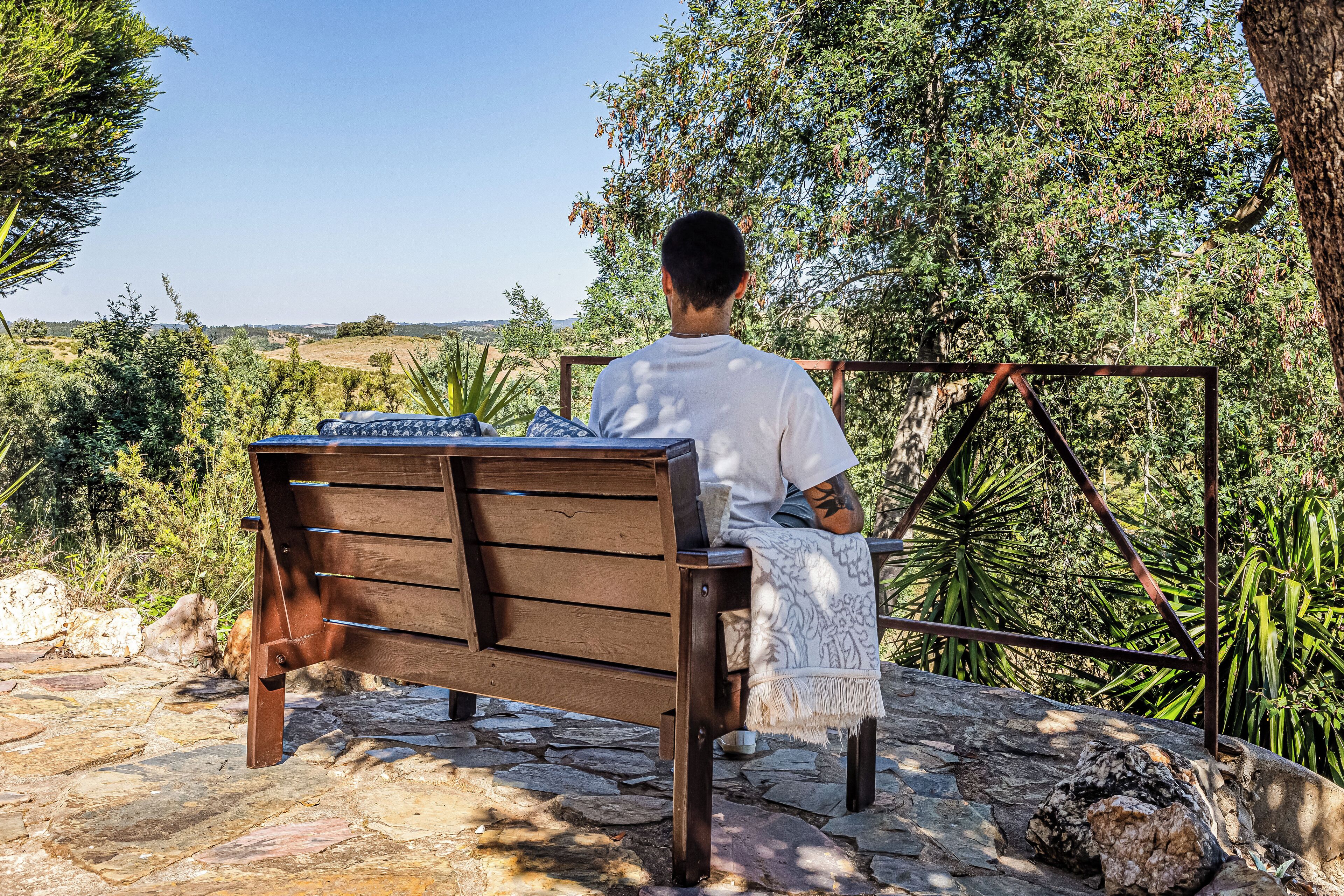 Une personne est assise sur un banc en bois à l’extérieur, d’où elle a une vue sur un paysage composé d’arbres et d’un sentier en pierre.