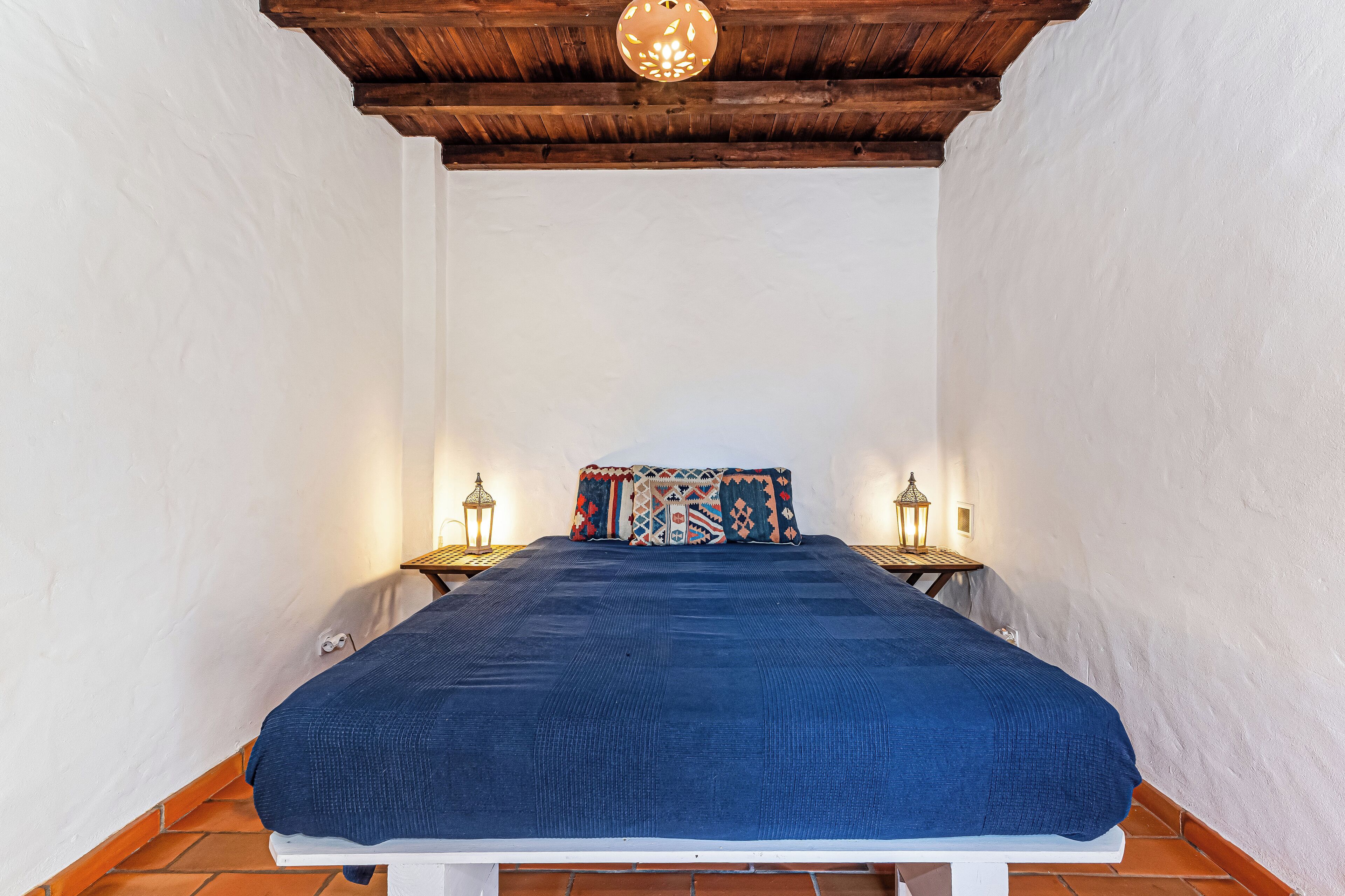 Une chambre avec un plafond en bois, des murs blancs, une couvre-lit bleue et deux tables de chevet surmontées de lampes de style lanterne.