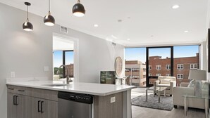 A modern kitchen with a dishwasher, a large island, and pendant lighting. There is a view of a brick building through large windows. - Global Luxury Suites Downtown Charleston (North Charleston)