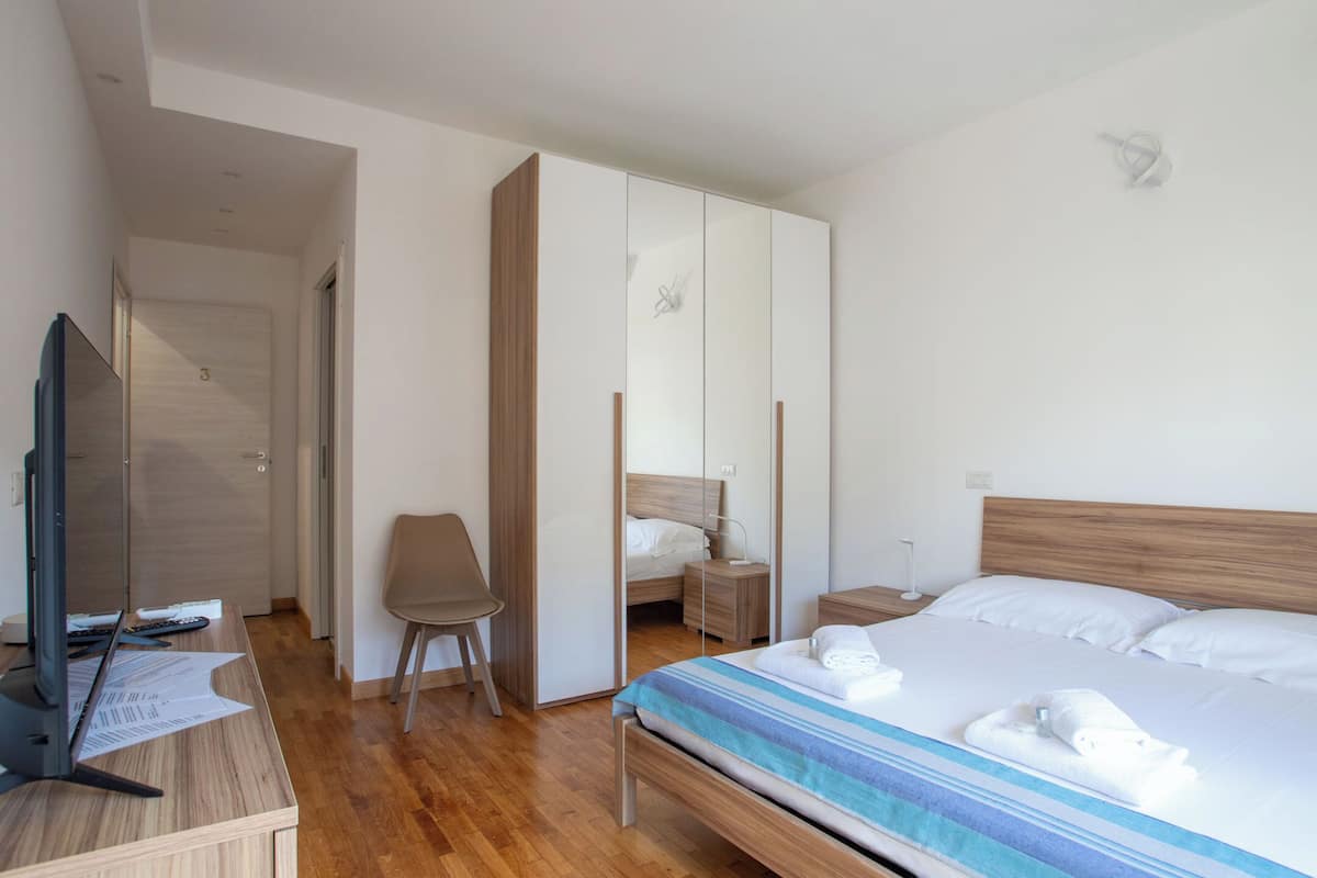 A hotel room with a wooden bed, a desk with a computer, a chair, a wardrobe, and a bedside table with a lamp.