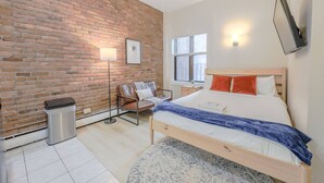 A bedroom with a brick wall, a bed with a wooden headboard, a floor lamp, a chair, a small trash bin, and a window with blinds. - Delightful South End Studio (Boston)