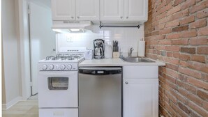 A compact kitchen with white appliances, a brick wall, and a door leading to another room. - Delightful South End Studio (Boston)