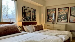Ein Schlafzimmer mit zwei Betten, einem hölzernen Schminktisch und einem Fenster mit Blick auf Bäume.