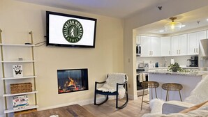 Ein Wohnzimmer mit Kamin, einem an der Wand befestigten Fernseher und einer durchgehenden Tür einsehbarer Küchenbereich.