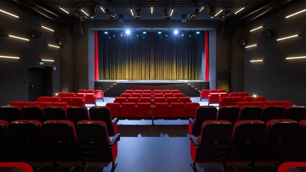 En teater med rækker af røde sæder, en scene med gardiner og loftsbelysning.