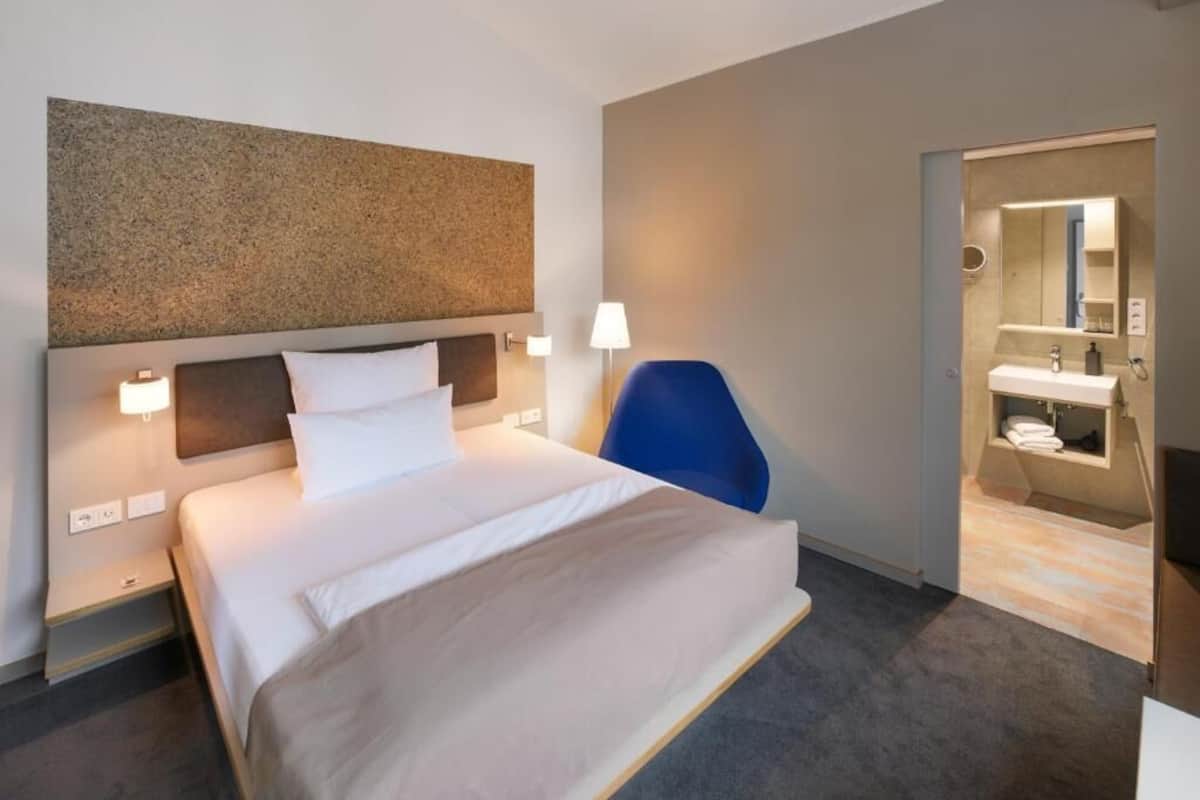 Hotellihuone, jossa on suuri sänky, yöpöydällä olevat lamput, sininen tuoli ja näkymä kylpyhuoneeseen.