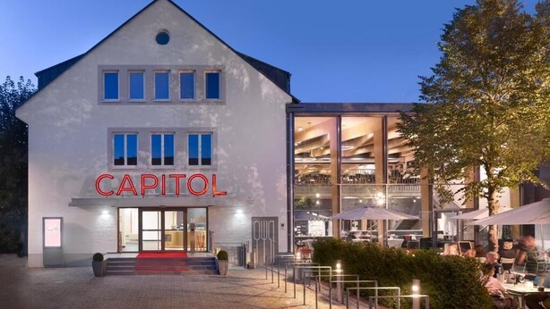 En byggnad med en framträdande skylt som visar ordet ”CAPITOL”, stora fönster och ett utomhusområde med sittplatser och parasoller.