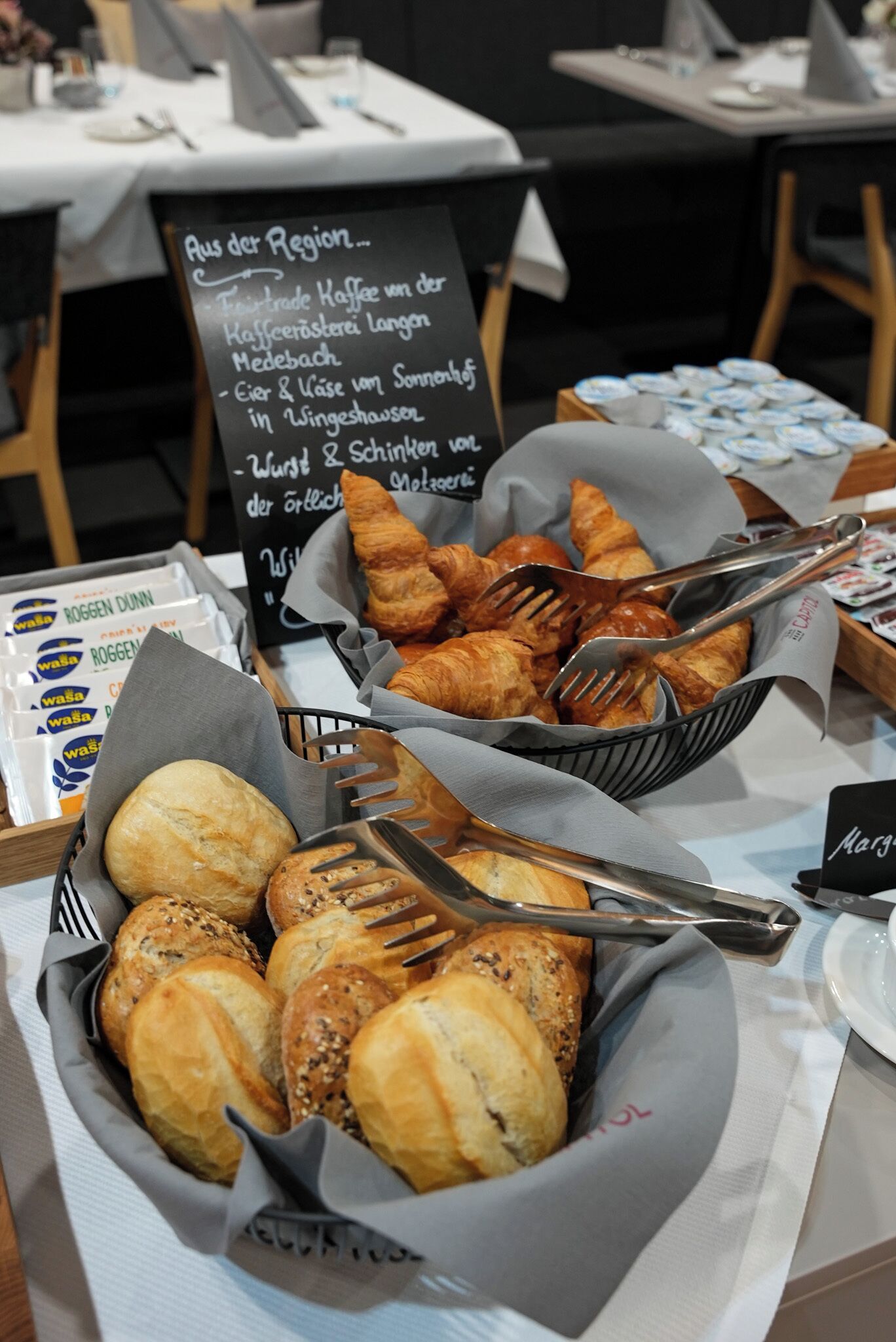 Ein reichhaltiges Buffet mit verschiedenen Brotsorten und Gebäck, darunter Croissants und Brötchen, mit einem Schild, das regionale Spezialitäten auflistet.