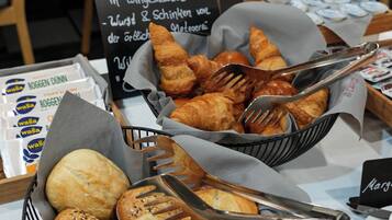 Ein reichhaltiges Buffet mit verschiedenen Brotsorten und Gebäck, darunter Croissants und Brötchen, mit einem Schild, das regionale Spezialitäten auflistet.