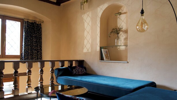 Een kamer met een gebogen blauwe bank, een kleine ronde tafel, een houten leuning en een hangende lamp.