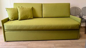 Un canapé vert avec deux coussins, dans une pièce dont le sol est recouvert de parquet et dont un des murs est blanc.