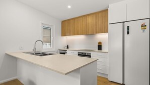A modern kitchen with a central island, wooden cabinets, a built-in oven, and a refrigerator with energy efficiency stickers. - Blenheim Breeze by Experience Jervis Bay (Vincentia)