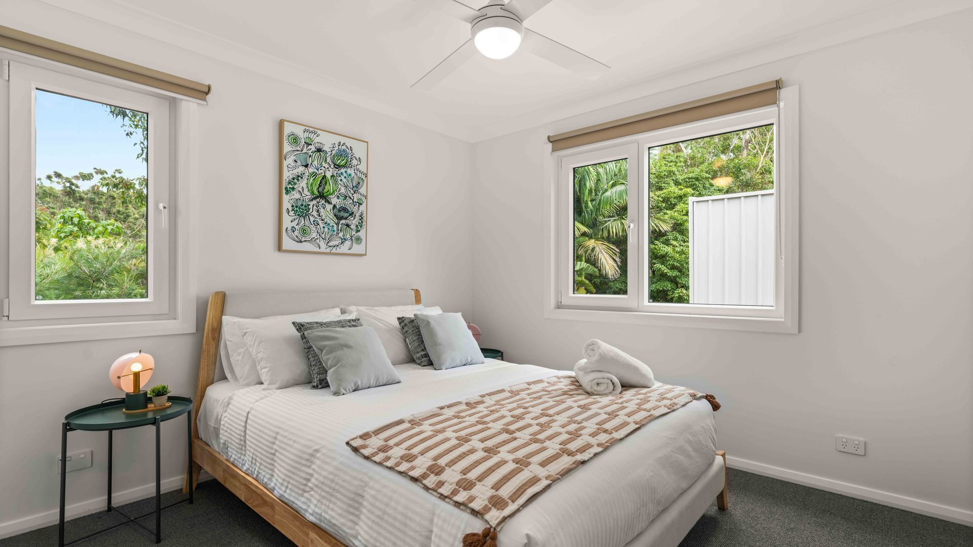 Une chambre à coucher comprenant un lit, une table de chevet, un ventilateur de plafond et deux fenêtres donnant sur un paysage verdoyant.