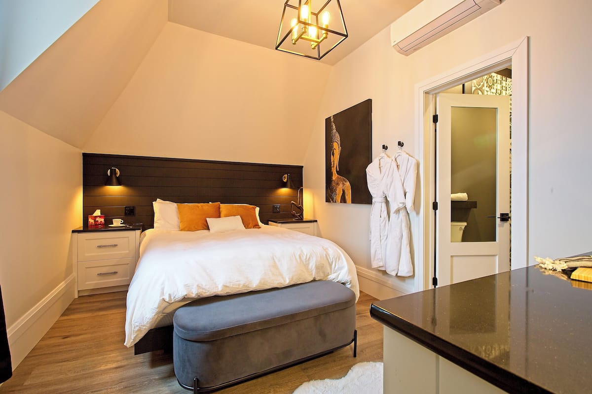 Een slaapkamer met een bed, nachtkastjes, een bankje, een kledingkast en een badkamer zichtbaar door een open deur.