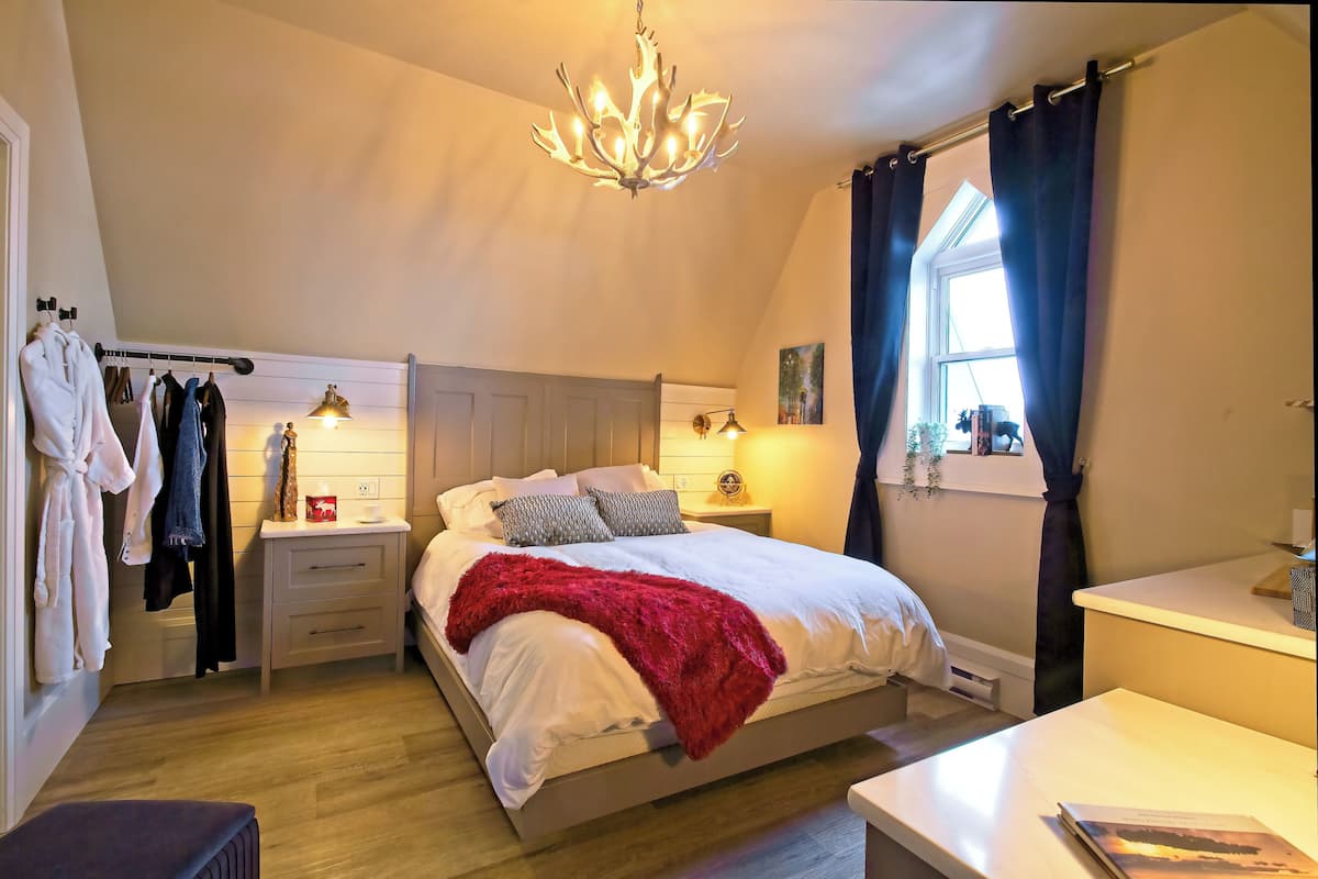 Een slaapkamer met een bed, nachtkastjes, een lamp, een kroonluchter, een raam met gordijnen en uitzicht op buiten.