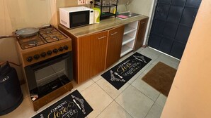 Een compacte keuken met een gasfornuis, oven, magnetron en houten kastjes. Er liggen twee zwarte deurmatten op de vloer met tekst.