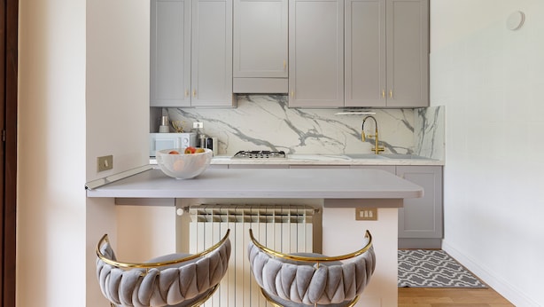 A modern kitchen with grey cabinets, a marble countertop, and two upholstered chairs with gold accents. - Raphael Stresa (Stresa)