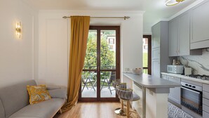 A modern kitchen with grey cabinets, a white countertop, and a built-in oven. There is a dining area with a grey sofa and two bar stools. A large window with a yellow curtain provides a view of the outdoors. - Raphael Stresa (Stresa)